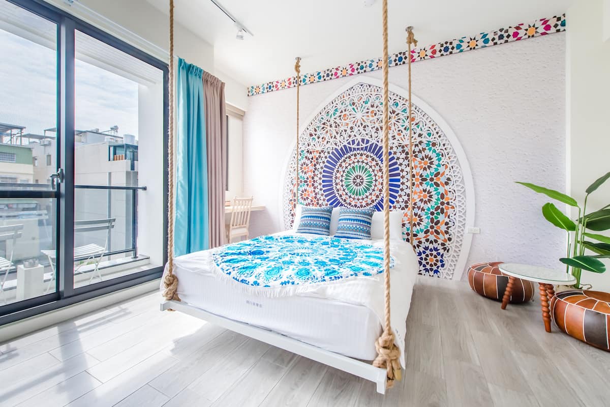 Une chambre à coucher avec un lit, une grande fenêtre, une vue sur des immeubles, une tapisserie murale et une plante.
