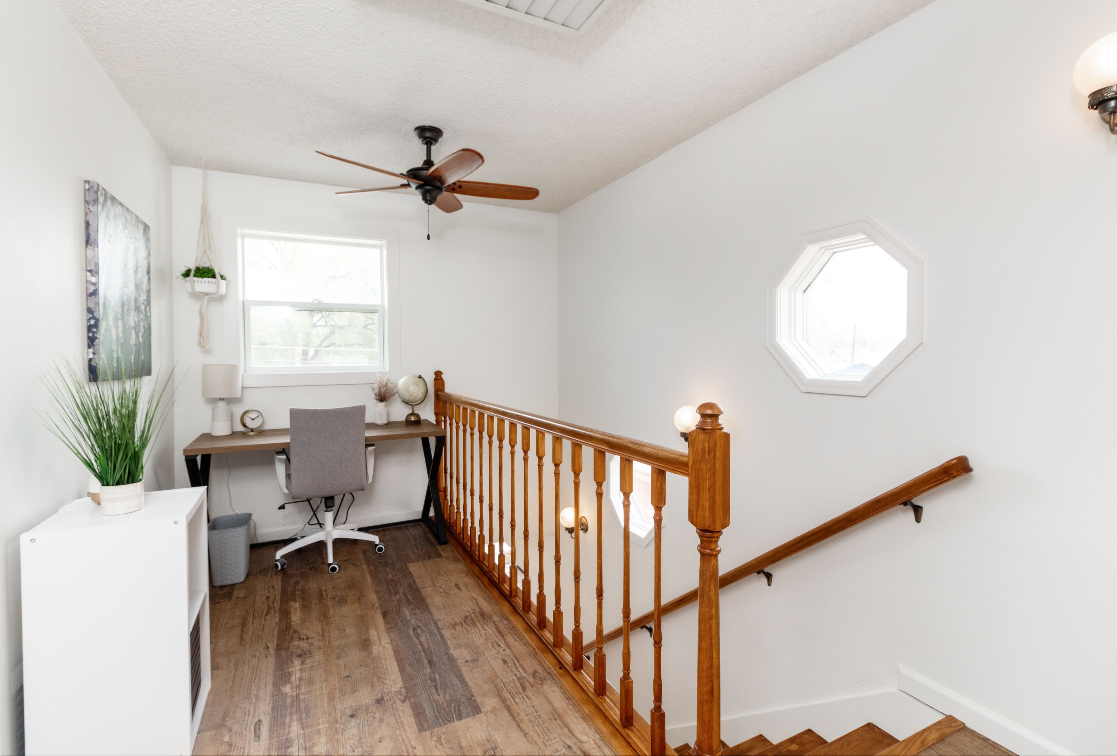 Una habitación con escalera de madera, un escritorio con ordenador, una silla y un ventilador de techo.