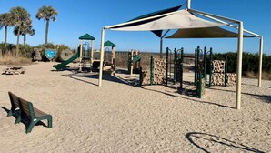 En lekplats vid stranden med ett skuggat område, en rutschkana och en bänk.