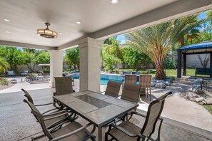 An outdoor patio with a dining table and chairs, surrounded by a pool and greenery.