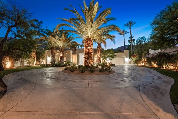 A well-lit driveway leading to a building with a modern architectural style, surrounded by palm trees and landscaped gardens.