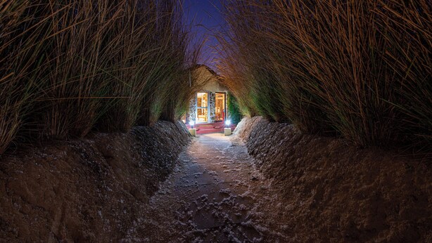A narrow pathway leading to a lit building surrounded by tall grass. - Jungle Cave Resort (Sawai Madhopur)