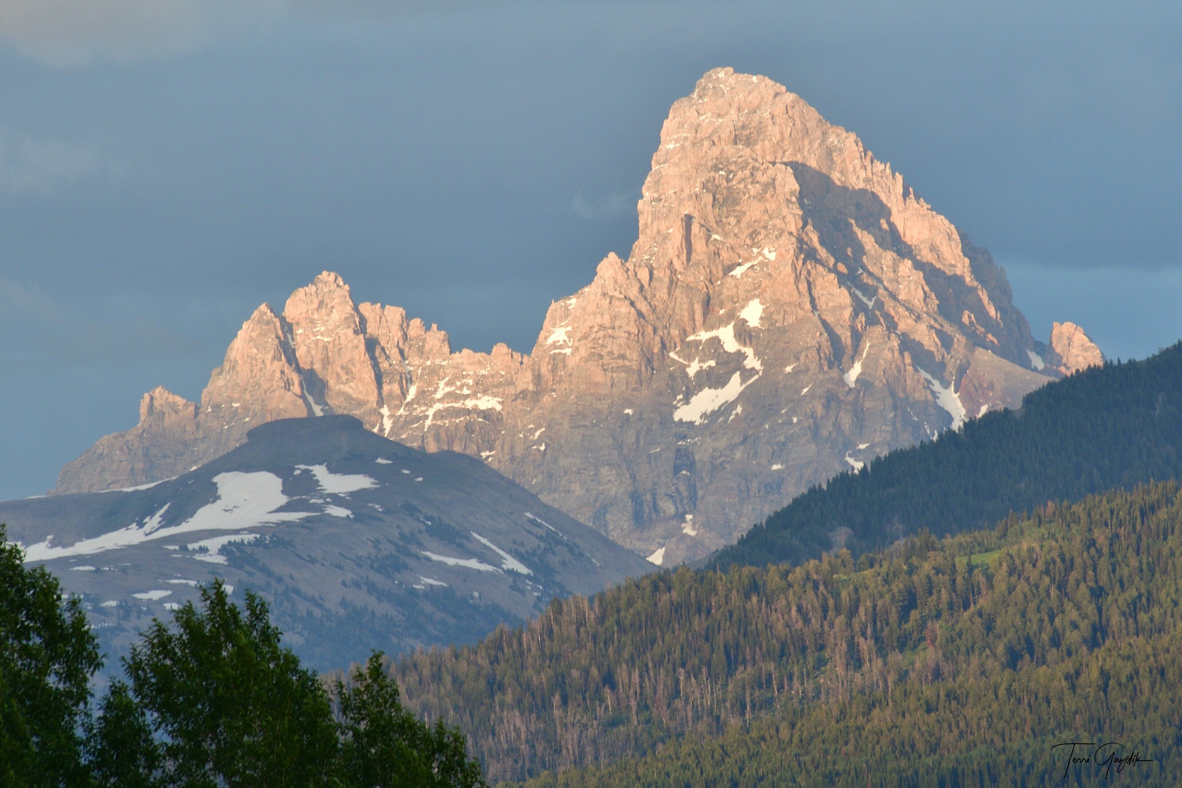 Paisagem montanhosa com um pico imponente, encostas arborizadas e um vale.