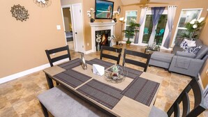 A dining area with a wooden table, black chairs, and a bench. There is a fireplace with a TV above it, and a grey sofa in the background. - Month Stays Available | Close to Hurlburt Field, Navarre & Okaloosa | Dogs Ok |Fenced Yard |Foosball (Mary Esther)