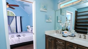A bedroom with a bed, a ceiling fan, and a window with curtains. The bathroom has a sink, mirror, and decorative items. - Month Stays Available | Close to Hurlburt Field, Navarre & Okaloosa | Dogs Ok |Fenced Yard |Foosball (Mary Esther)