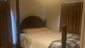 A bedroom with a bed, a wooden headboard, a quilted bedspread, a plaid blanket, a nightstand, and a stained glass light fixture.