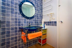 Ein Badezimmer mit blauen Fliesen, einem runden Spiegel, einem Waschbecken aus Glas und einem Handtuchhalter mit einem orangefarbenen Handtuch.