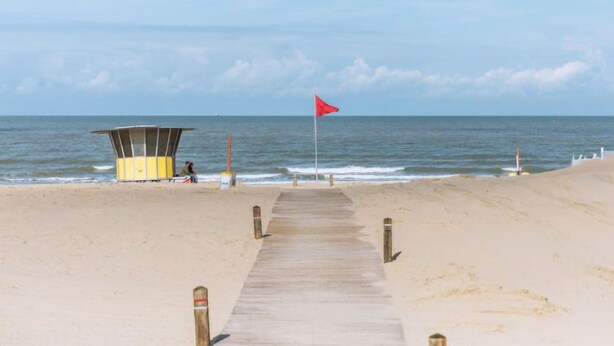 Ein Strand mit einer Holzpromenade, die zu einem Rettungsturm führt, und eine rote Flagge.