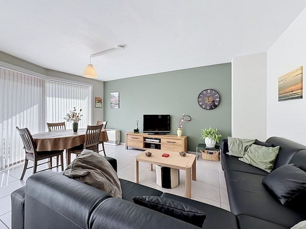 A modern living room with a black leather sofa, a dining table with chairs, a TV, and a clock on the wall.