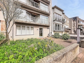A multi-story residential building with balconies, a brick facade, and a garden area with various plants.
