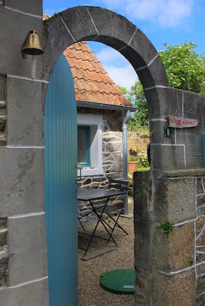 A stone archway leading to a small building with a tiled roof, a bell mounted on the wall, and a sign reading "L'ABRI OU GUNDY".