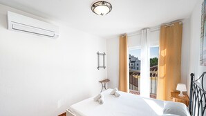 Ein Schlafzimmer mit einem Bett, einem kleinen Nachttisch mit Lampe, einem Fenster mit Vorhängen und einer Klimaanlage.