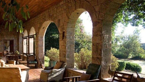 Un área de estar al aire libre con techo, arcos de piedra, muebles de madera y vistas al verdor.