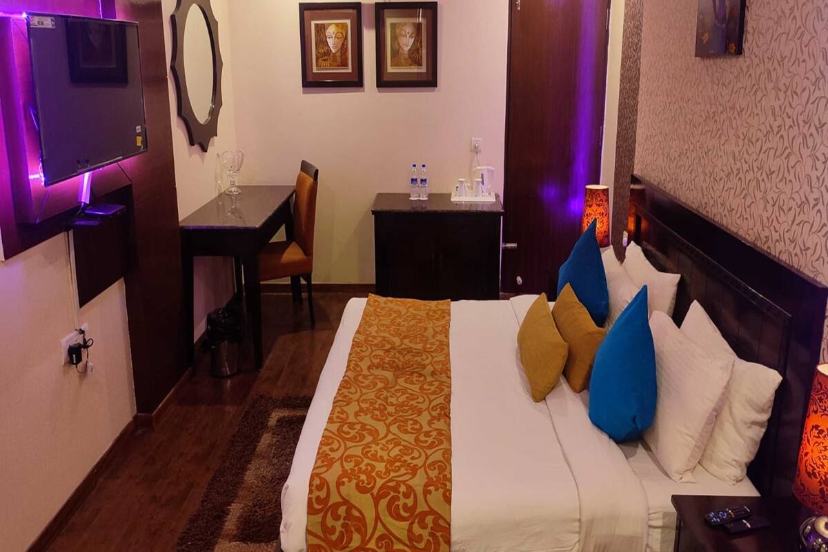A hotel room with a bed, bedside table, TV, desk, chair, and artwork on the walls.