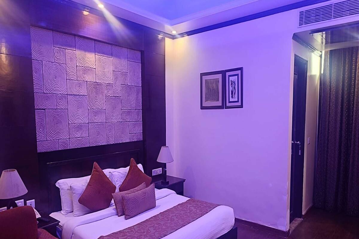 A modern hotel room with a large bed, a headboard with a geometric pattern, two bedside tables with lamps, a red armchair, and a door leading to another room.