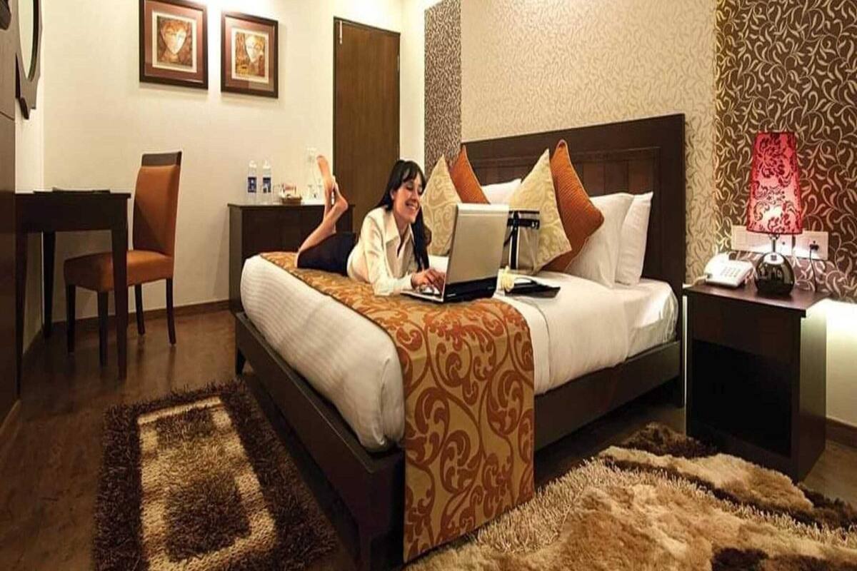A hotel room with a bed, a desk, a chair, a lamp, and a picture frame on the wall.