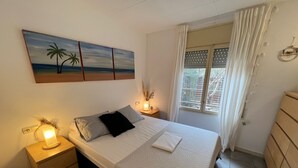 Et pænt møbleret soveværelse med seng, sengeborde, et vindue med gardiner og et maleri med et strandmotiv på væggen.