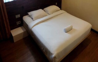 Una cama bien hecha con sábanas blancas y una toalla enrollada, un cabecero de madera y una mesita de noche con una lámpara.