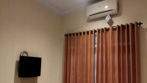 A room with beige walls, a mounted flat-screen TV, a window with vertical blinds, and an air conditioning unit above the window. - Griya Joglo Kaliurang (Ngaglik)