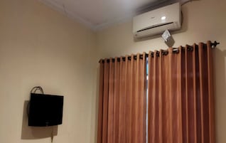 Una habitación con paredes color beige, un televisor de pantalla plana montado, una ventana con persianas verticales y un equipo de aire acondicionado sobre la ventana.