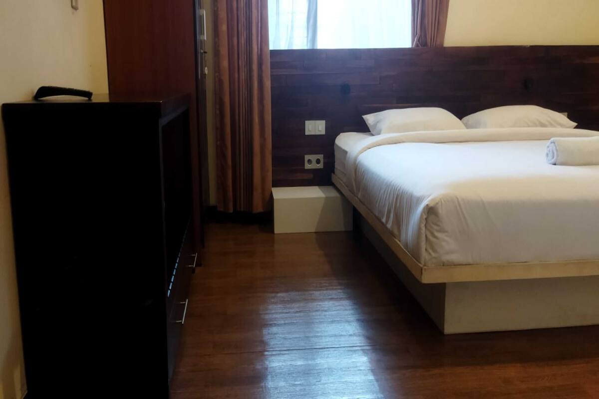 Una habitación de hotel con cabecera de madera, cama con ropa blanca, una mesita de noche pequeña y un armario de color oscuro.