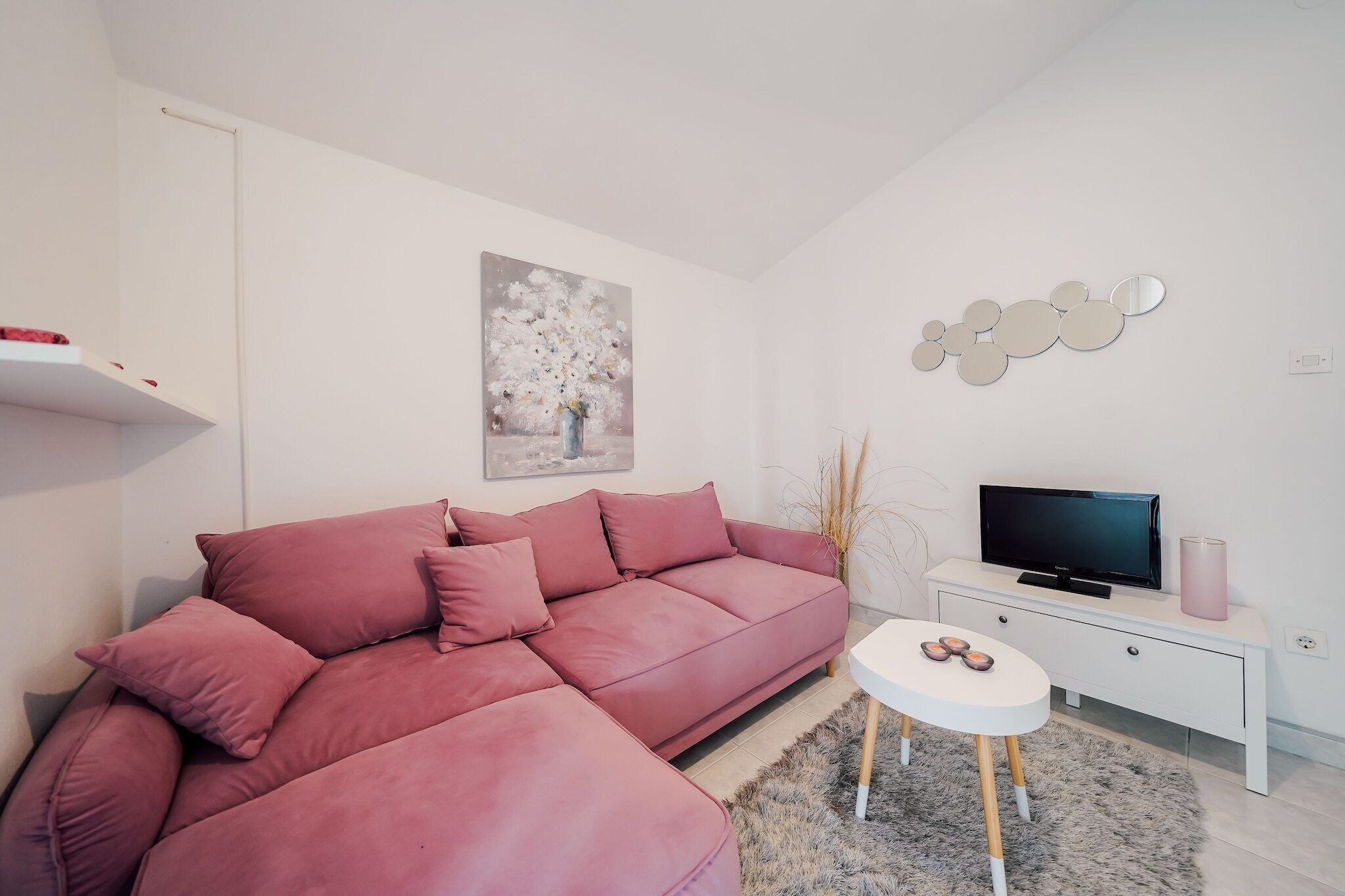 A modern living room with a pink sectional sofa, a flat-screen TV on a white console table, a small white side table, and a painting of flowers on the wall.