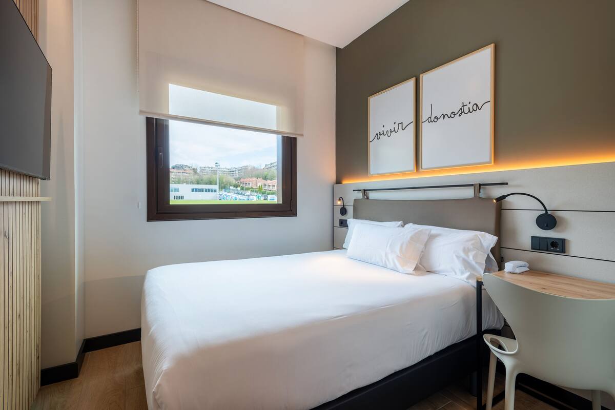 A modern hotel room with a large bed, a desk, and a view of a cityscape.