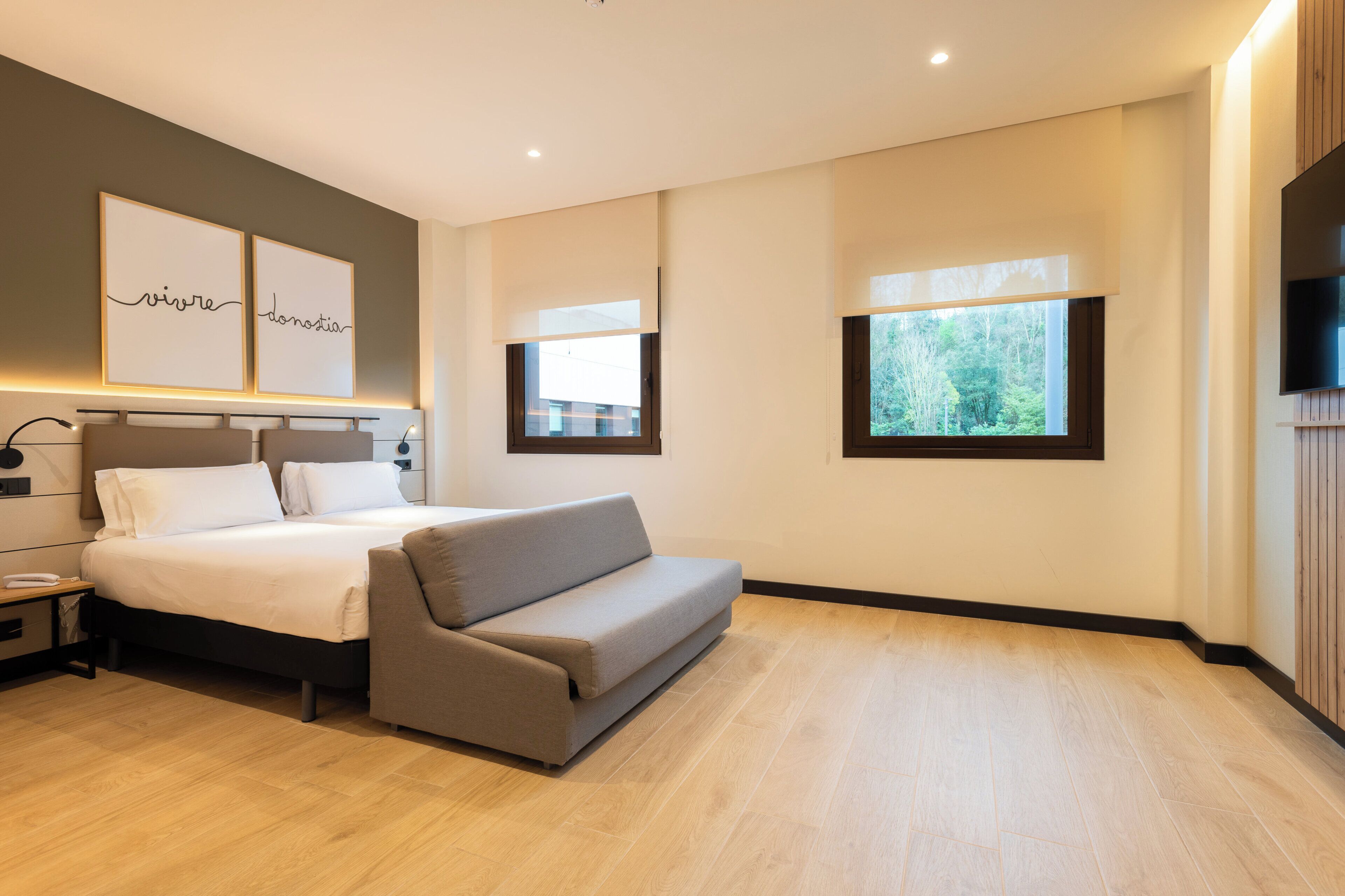 A modern hotel room with a bed, a sofa, a TV, and artwork on the wall.