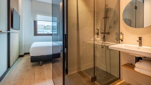 A modern hotel room with a glass-enclosed shower, a large bed, a mounted TV, and a view of the city through a window. - Leonardo Hotel San Sebastián (San Sebastián)