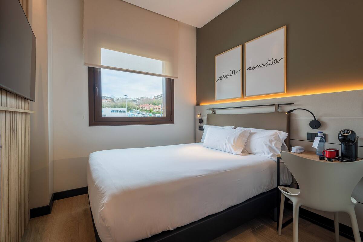 A hotel room with a bed, a desk with a chair, a window with a view, and two framed pictures on the wall.