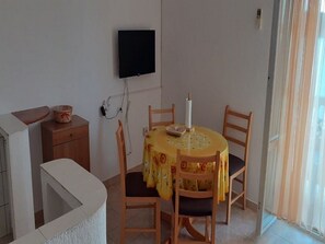 A dining area with a round table covered by a yellow tablecloth, two wooden chairs, a television mounted on the wall, and a small wooden cabinet with a basket on top.