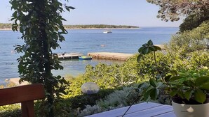A wooden table with a potted plant, surrounded by greenery and overlooking a calm body of water with boats.