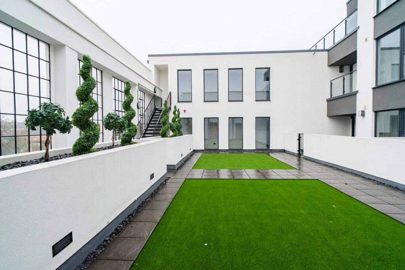 A modern building with a courtyard featuring artificial grass and neatly trimmed topiary plants.