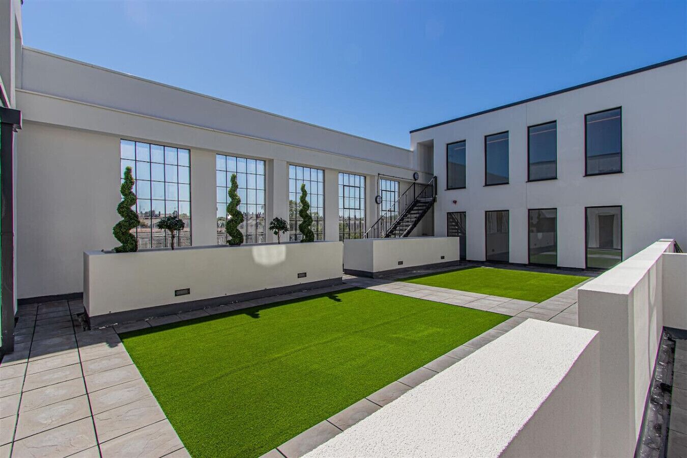 A rooftop terrace with artificial grass, a white building with large windows, and a staircase leading to an upper level.