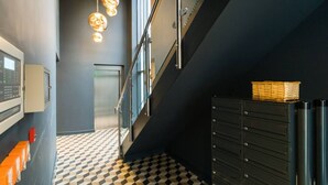 A modern hallway with a checkered floor, a staircase, a metal railing, and a black filing cabinet with a basket on top. - Exquisite 2 Bed Duplex Apt | Secure Parking | (Penarth)