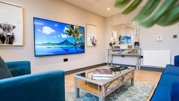 A modern living room with a large TV, blue sofas, a coffee table, and a decorative mirror. - Exquisite 2 Bed Duplex Apt | Secure Parking | (Penarth)