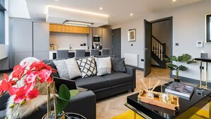 A modern living room with a grey sofa, a coffee table, and a plant. There is a kitchen area in the background with a microwave and a stove. - Luxe 2 Bed Duplex Apt w/Balcony, 3 Bathrooms, Free Parking (Penarth)
