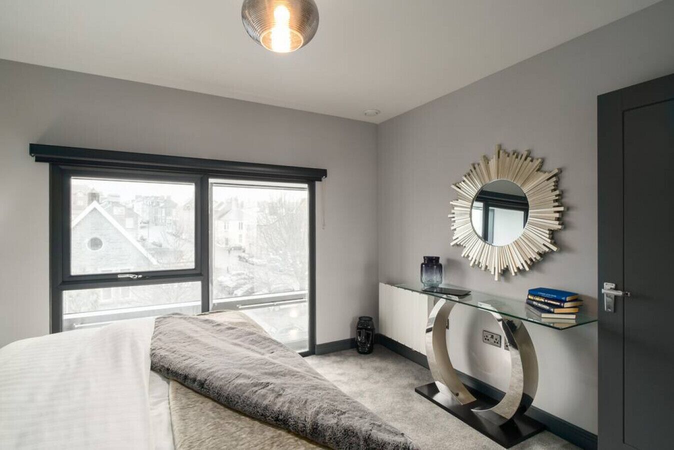 A bedroom with a bed, a glass desk, a mirror, and a view through large windows.