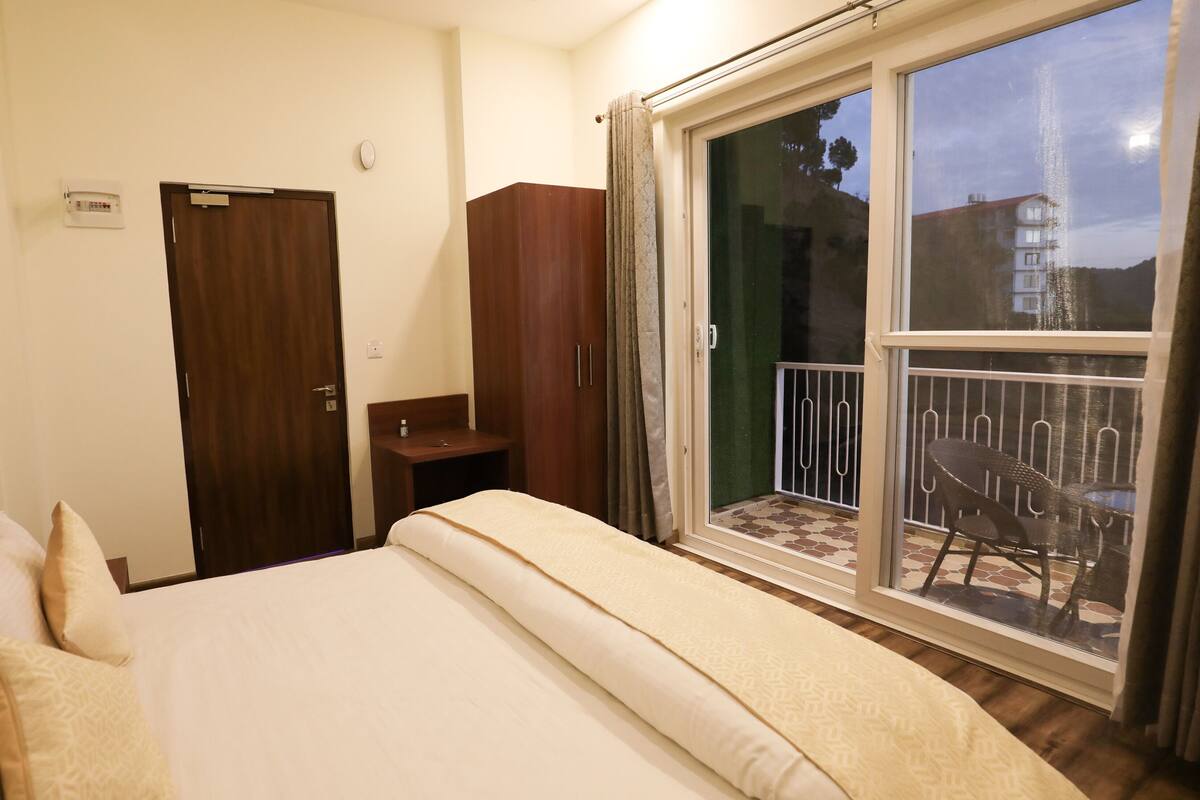 A hotel room with a bed, a wardrobe, a small table, a window with a view of the outside, and a balcony with chairs.