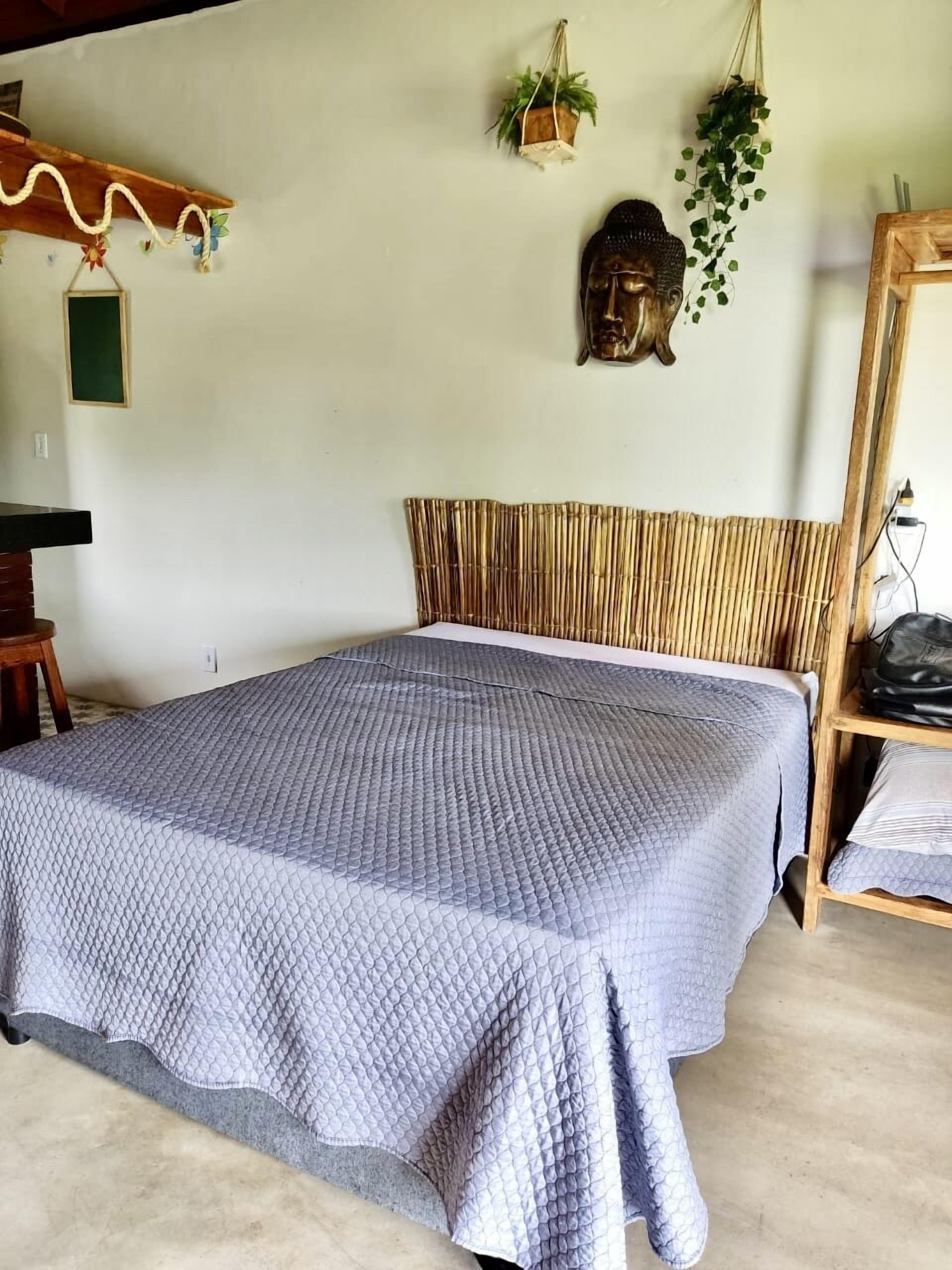 Una habitación con cama, cabecera de madera, estatua de Buda montada en la pared y plantas colgantes.