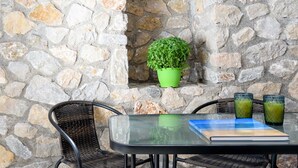 Un mur de pierres avec un pot de plante verte, une chaise en osier, une table avec un livre et deux verres contenant une boisson verte.