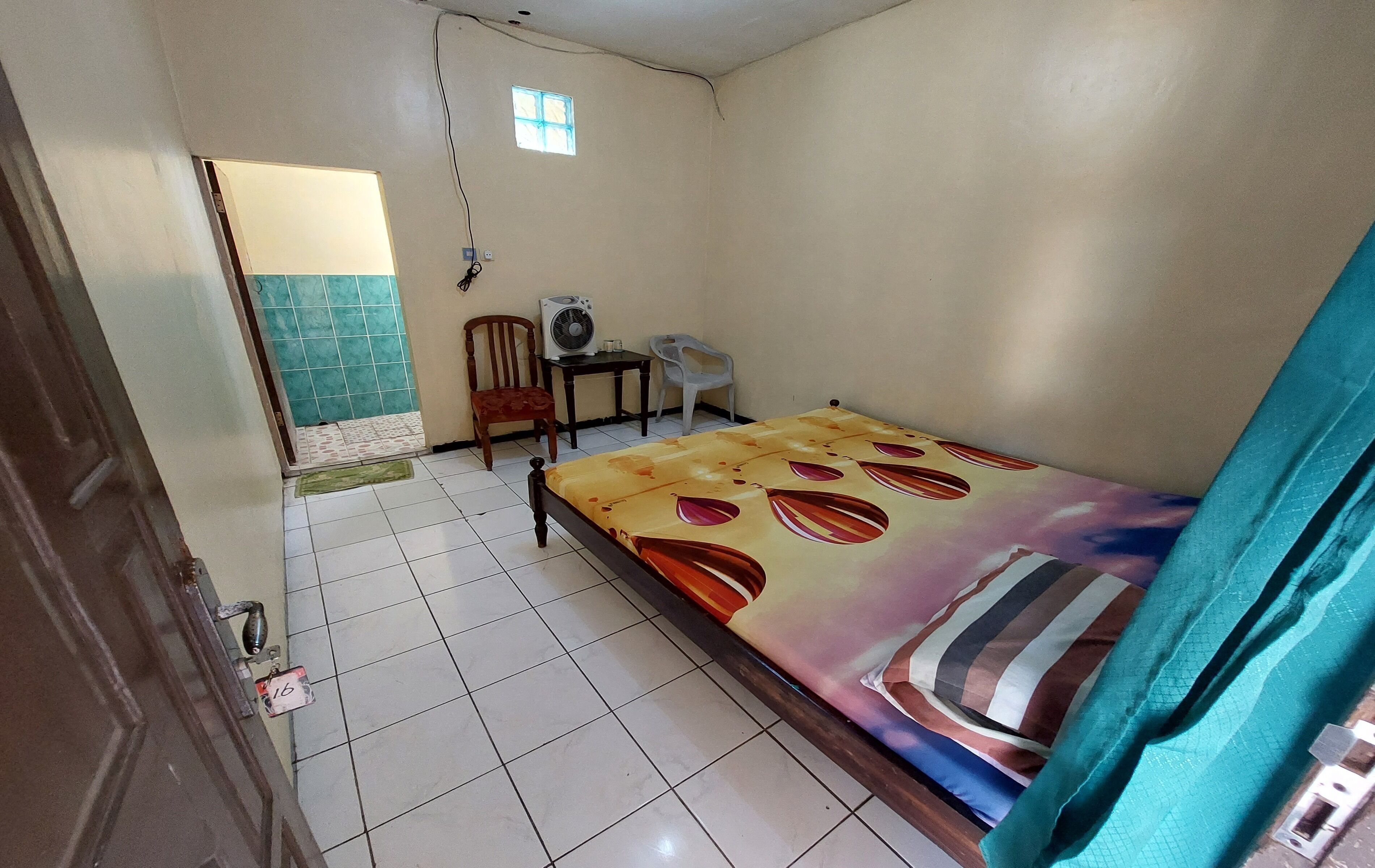 A small, clean room with a bed, a chair, a table, and a fan.