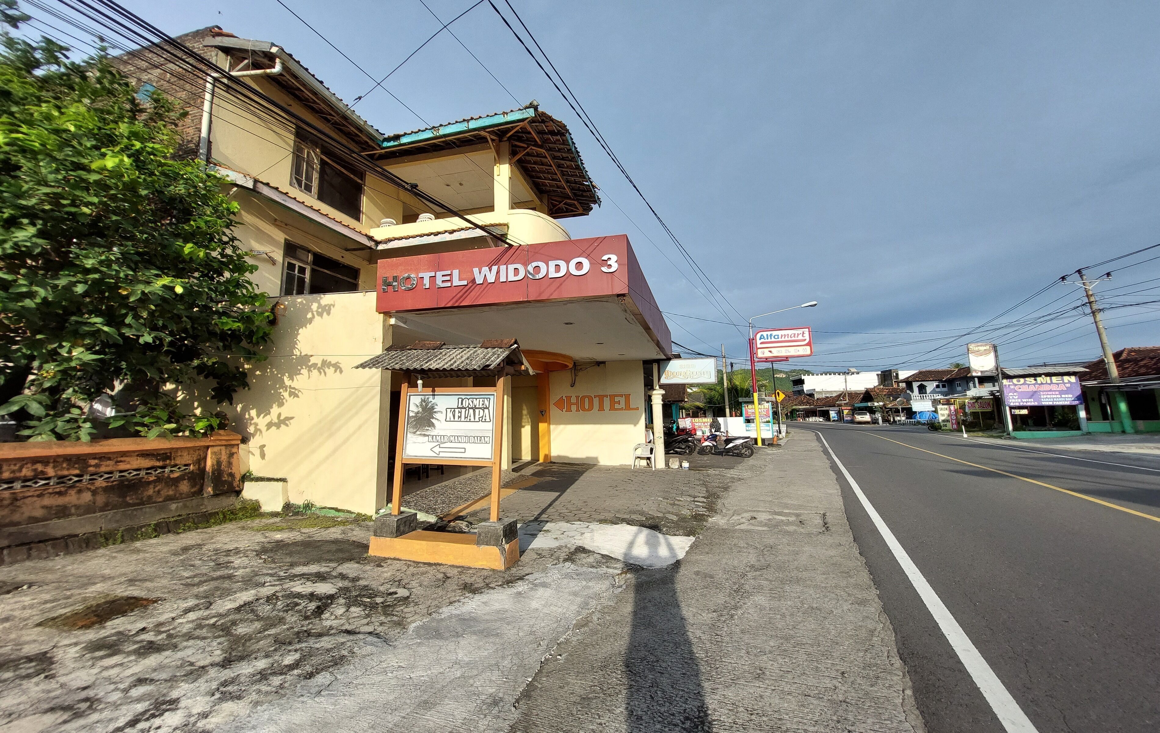 A hotel named 'Hotel Widodo 3' with a signboard, a small outdoor seating area, and a road in the background.