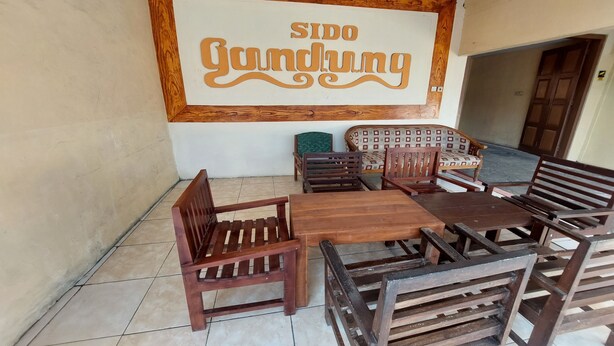 Un ensemble de salle à manger en bois, composé d’une table et de chaises, se trouve dans une pièce dont le sol est carrelé et sur un des murs est accrochée une enseigne portant l’inscription « SIDO Gundung ».