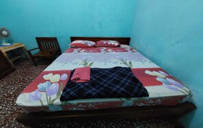 A bed with a floral bedspread, a wooden chair, and a small table with a fan.