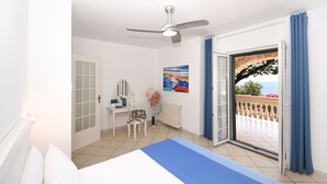 Un dormitorio con cama, escritorio, silla, ventilador de techo, ventana con cortinas y vista al balcón exterior.