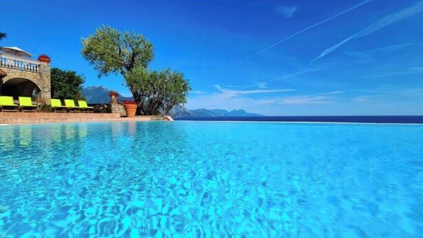Una piscina azul cristalina con un edificio de piedra y sillones de descanso verdes a la izquierda.