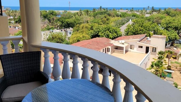 A balcony with a glass table and wicker chair, overlooking a coastal area with buildings and greenery.