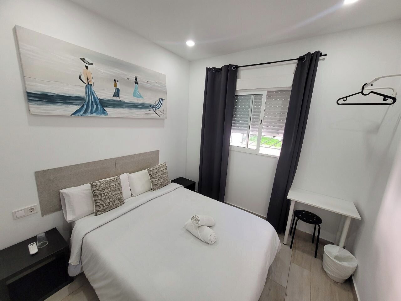 Una habitación de hotel moderna con una cama grande, una mesita de noche, un escritorio pequeño, un perchero y una ventana con cortinas.