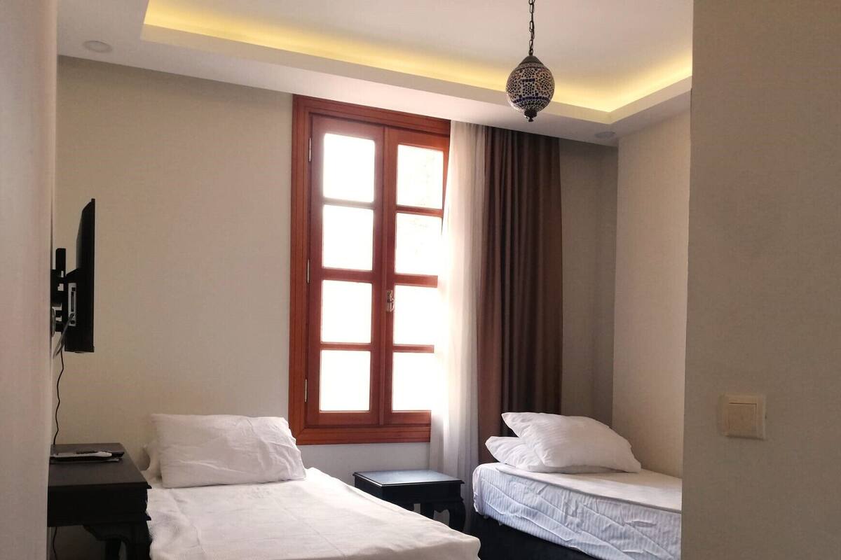 A hotel room with two beds, a desk, a TV, a window with curtains, and a hanging lamp.